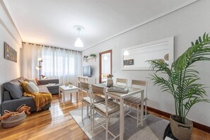Ein modernes Wohnzimmer mit Sofa, Couchtisch, Esstisch und Stühlen. Es befindet sich ein Fernseher, eine Pflanze und Kunstwerke an den Wänden.