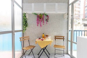 Un balcon avec une table dressée pour deux personnes, des chaises, une nappe à carreaux et des plantes suspendues.