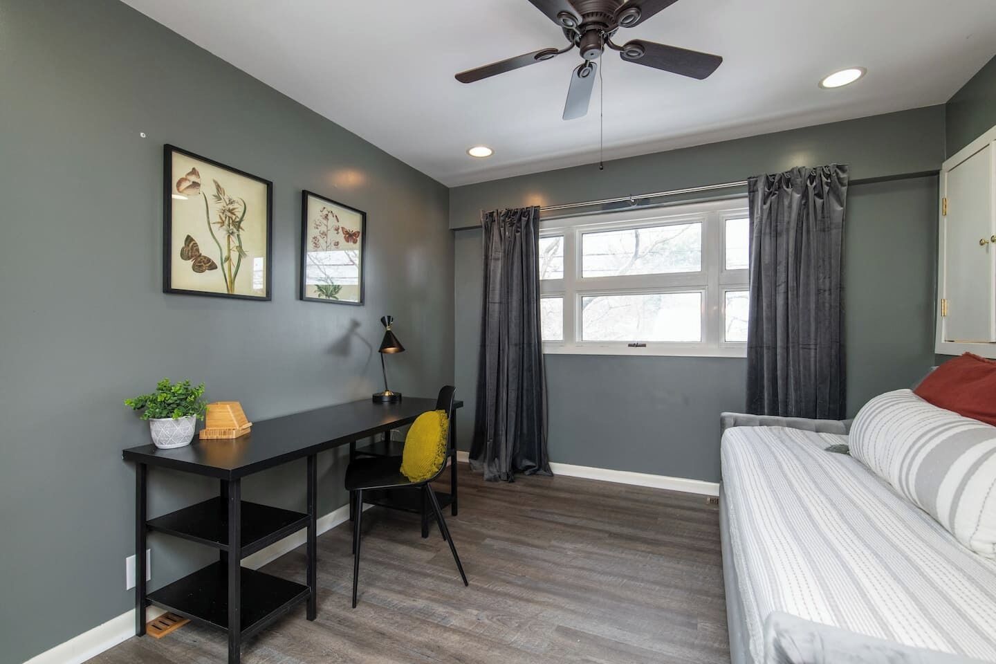 A bedroom with a bed, a desk, a chair, a ceiling fan, and a window with curtains.