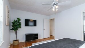 Quarto com cama, televisão, planta, ventoinha no teto e porta que dá para outro cômodo.