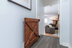 Ein Flur mit einer Holztür im Bauernhausstil, einem grauen Teppich und einem Wohnzimmer mit grauem Sofa und einem Kamin aus Backsteinen im Hintergrund.