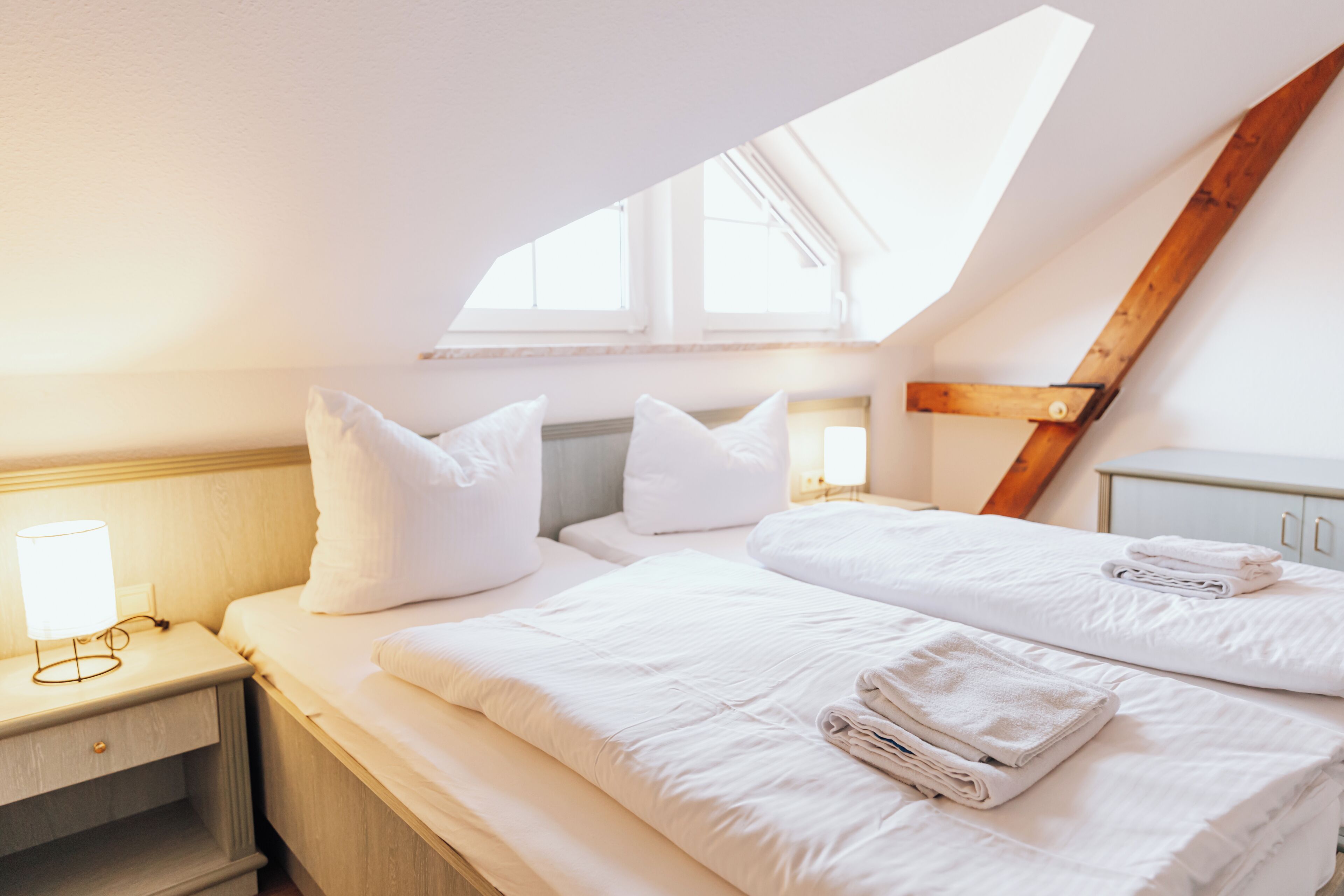 A bedroom with a sloped ceiling, a bed with white linens, a bedside table with a lamp, and a window providing natural light.