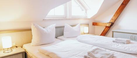 A bedroom with a sloped ceiling, a bed with white linens, a bedside table with a lamp, and a window providing natural light.