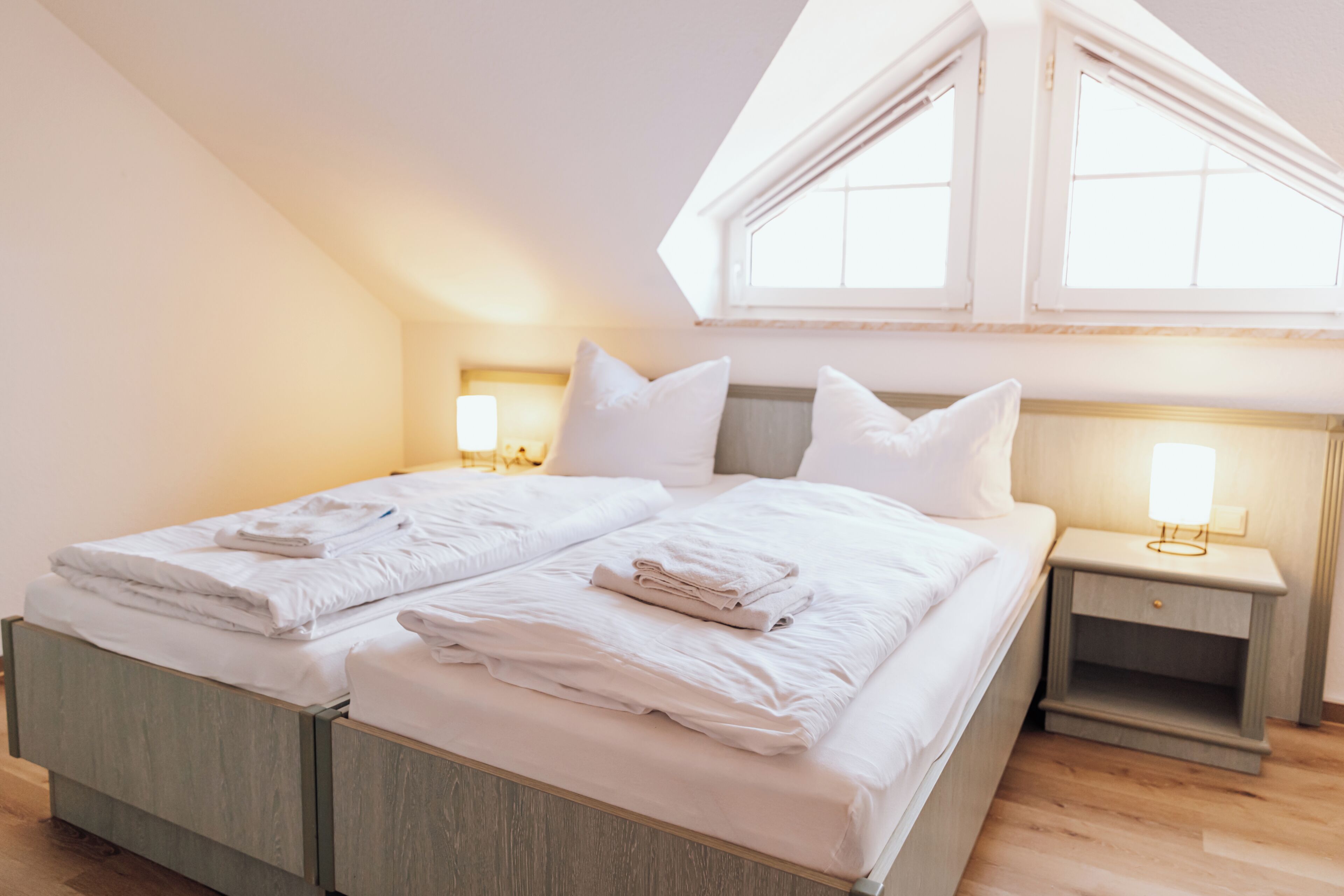 A twin bed with two pillows and folded towels, a bedside table with a lamp, and a window with a view of the sky.