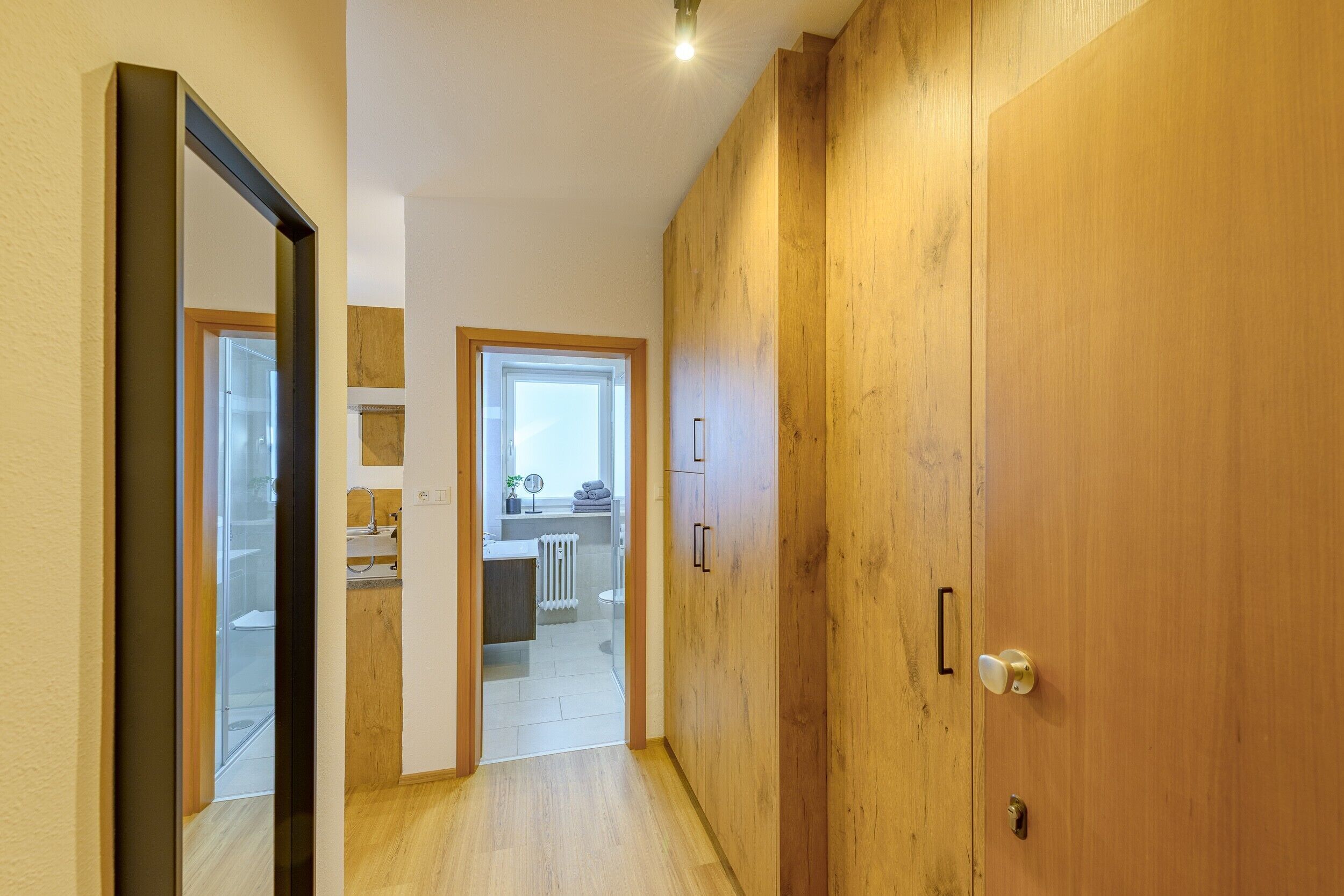 Ein Flur, der zu einem Badezimmer mit einem Holzschrank, einem Spiegel und einem Fenster führt.