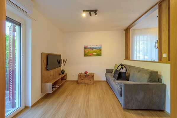 Un salon avec un canapé gris, un meuble TV en bois, un tableau accroché au mur et une fenêtre avec des stores.