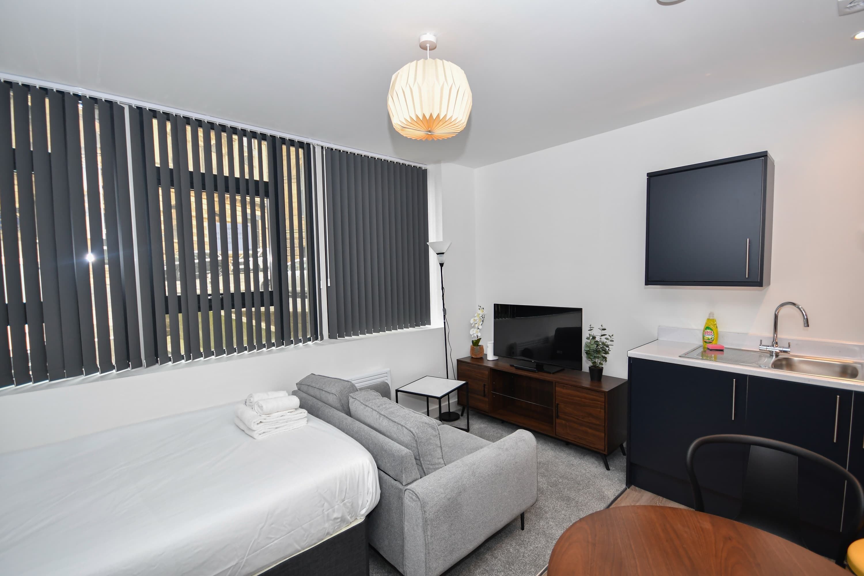 Ein modernes Schlafzimmer mit einem Bett, einem grauen Sofa, einem Fernseher, einem kleinen Tisch und einem Fenster mit Jalousien.