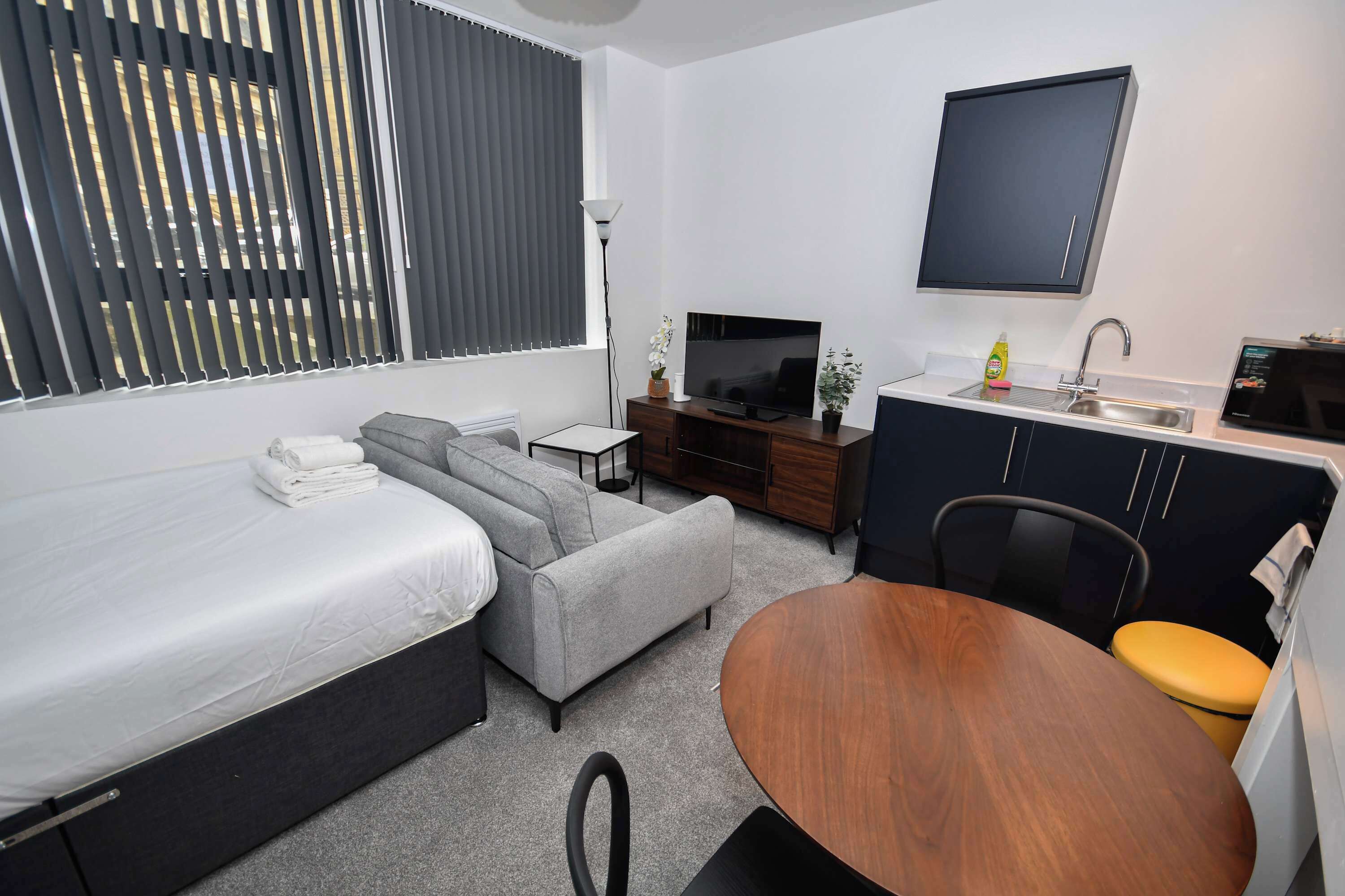 A compact hotel room with a bed, sofa, TV, kitchenette, and a dining table.