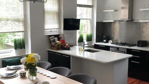 Eine moderne Küche mit einer zentralen Kochinsel, einem Essbereich mit Holztisch und einem großen Fenster mit Jalousien.