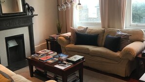 Ein Wohnzimmer mit Kamin, einer Couch, einem Couchtisch mit Büchern und einem Fenster mit Vorhängen.