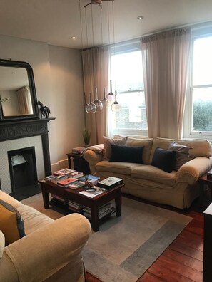 Ein Wohnzimmer mit Kamin, einer Couch, einem Couchtisch mit Büchern und einem Fenster mit Vorhängen.