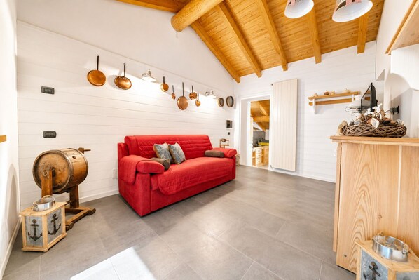 A spacious room with a red sofa, wooden ceiling beams, and a kitchen area visible through an open door.