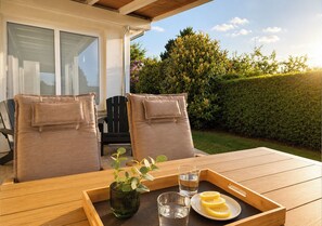 An outdoor seating area with two cushioned chairs, a wooden table, a tray with a glass of water, a plate with lemon slices, and a vase with green foliage.