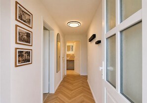 A hallway with framed pictures on the wall, a wooden herringbone floor, and a door leading to a bathroom.