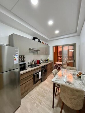 A modern kitchen with a built-in oven, microwave, and dishwasher, a dining area with a marble table, and a view into the living area. - Bel Appartement en Résidence et Piscine (Marrakech)