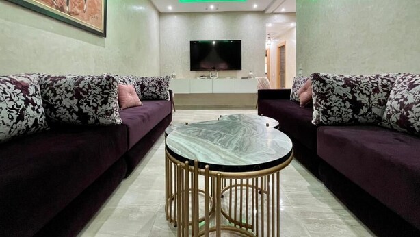 A living room with a sofa set, a round table, a flat-screen TV, and a green ceiling light. - Bel Appartement en Résidence et Piscine (Marrakech)