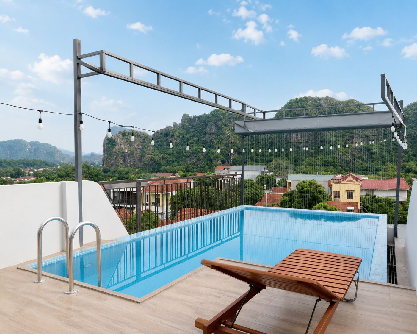 A rooftop swimming pool with a wooden lounge chair, metal railings, and a view of a mountainous landscape.