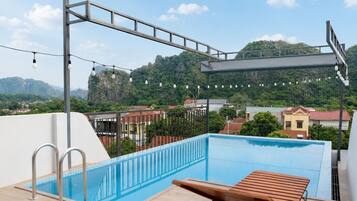 A rooftop swimming pool with a wooden lounge chair, metal railings, and a view of a mountainous landscape.