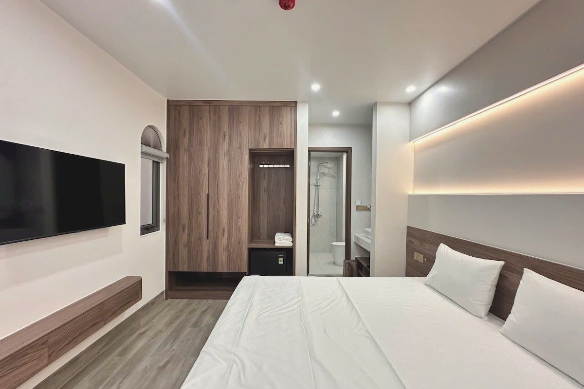 A modern hotel room with a large bed, a flat-screen TV mounted on the wall, a wooden wardrobe, and a walk-in closet.