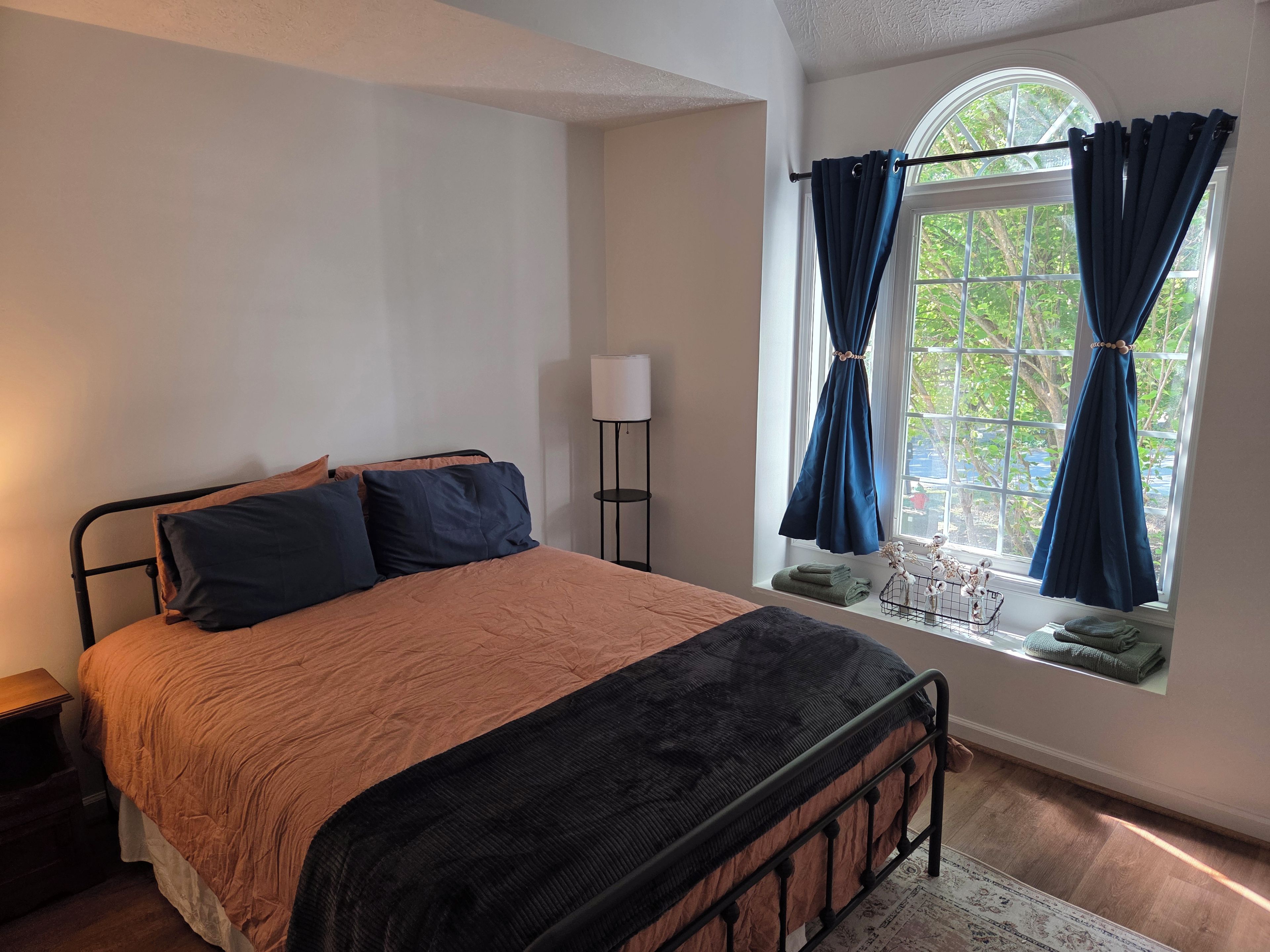 Une chambre à coucher avec un lit, une fenêtre avec des rideaux, une lampe de chevet et une petite table avec des serviettes.