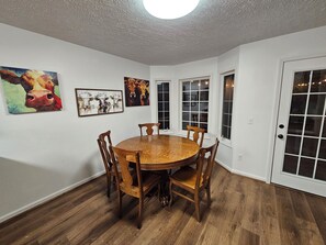 A dining room with a round wooden table and six chairs, white walls, and three paintings.