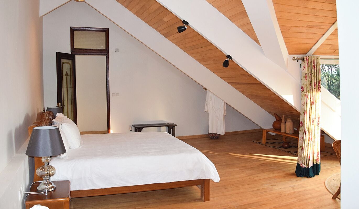 A bedroom with a sloped ceiling, a bed, a bedside table with a lamp, a mirror, and a window with curtains.