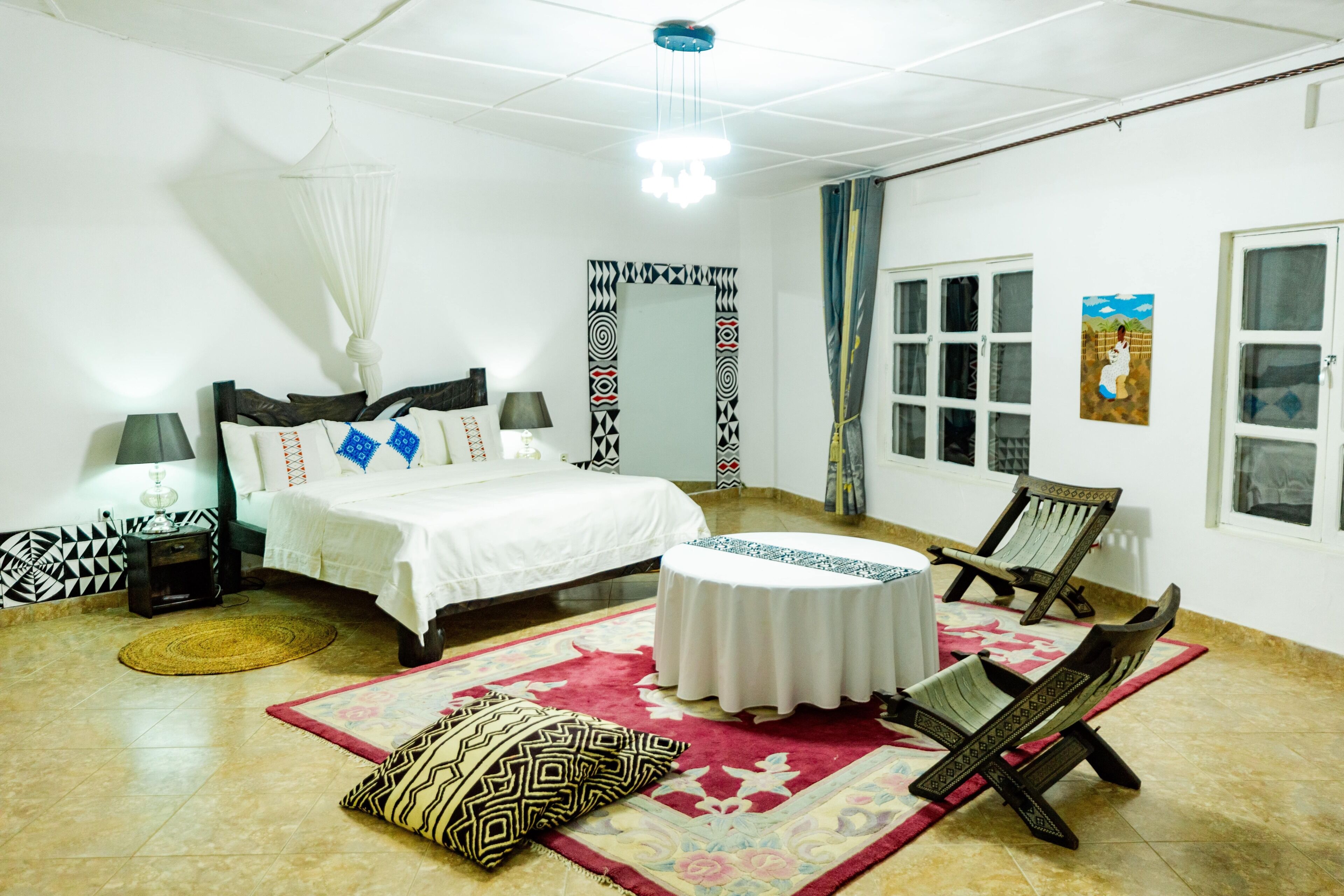 Un dormitorio con cama, mesitas de noche, una silla, una mesa redonda y una ventana.