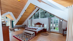 Un dormitorio con techo inclinado, una cama grande, un banco y vistas a zonas verdes a través de ventanas.