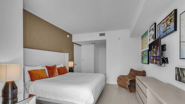 A modern hotel room with a large bed, a bedside table, a lamp, a chair, and a wall with framed artwork. - Oceanview Highrise - Amenities (Hollywood)