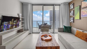A modern living room with a flat-screen TV, a wooden coffee table with remote controls, a sofa with cushions, and a balcony with a city view. - Oceanview Highrise - Amenities (Hollywood)