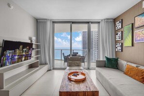 Ein modernes Wohnzimmer mit Fernseher, Couch mit Kissen, Couchtisch aus Holz mit Fernbedienungen und einem Balkon mit Blick auf die Stadt.