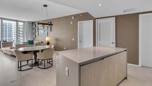 A modern kitchen with a central island, dining area with chairs, and a view of the cityscape through large windows. - Oceanview Highrise - Amenities (Hollywood)