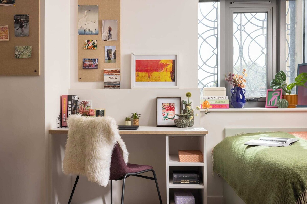 A room with a desk, a chair, a bed, a window with a view, and various items on the desk and shelves.