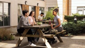An outdoor seating area with wooden tables and benches, surrounded by greenery and a building with large windows. - Student Only Cathedral Park (Bristol)
