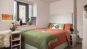 A bedroom with a bed, a nightstand, a window with plants, and a shelf with books and decorative items. - Student Only Cathedral Park (Bristol)