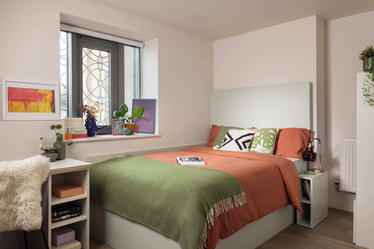 A bedroom with a bed, a nightstand, a window with plants, and a shelf with books and decorative items.