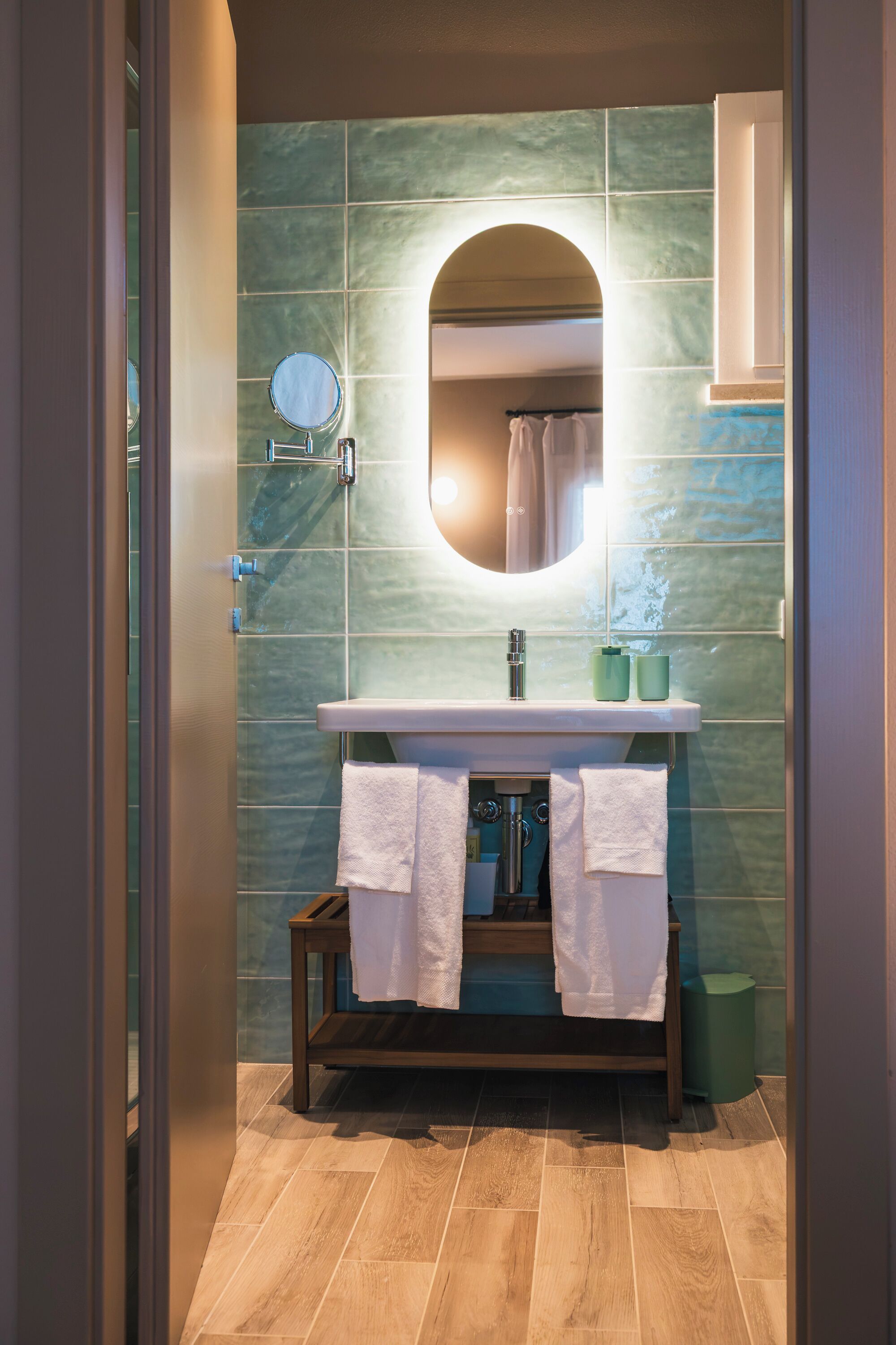 A bathroom with a wooden vanity, a round mirror, and a green tiled wall.