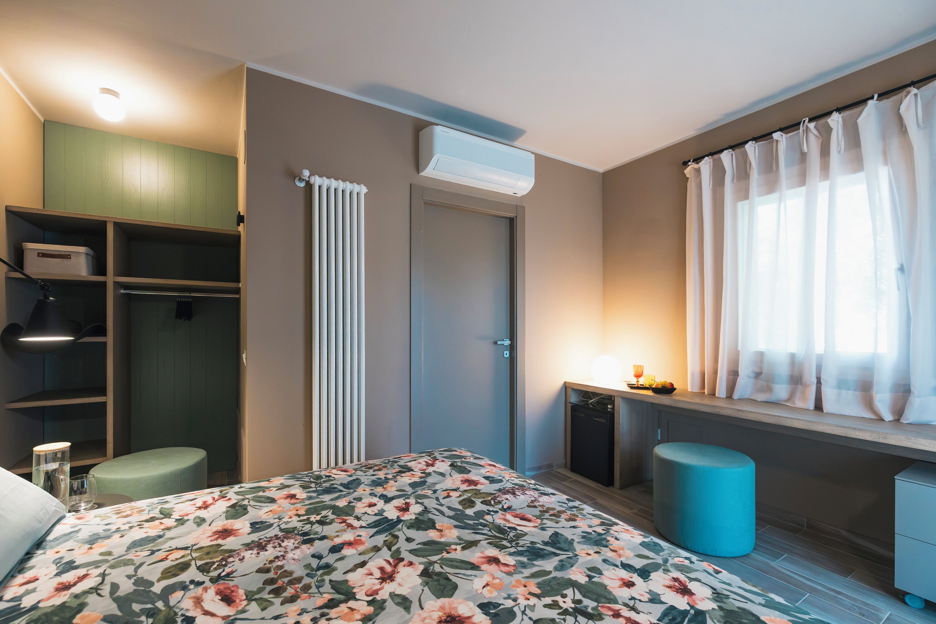 Un dormitorio moderno con cama, armario, radiador, ventana con cortinas y escritorio con lámpara.
