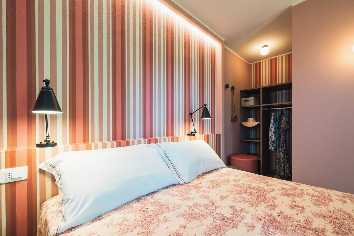 Ein Schlafzimmer mit einem Bett, zwei Nachttischlampen, einer gestreiften Wand, einem Kleiderschrank und einer Deckenleuchte.