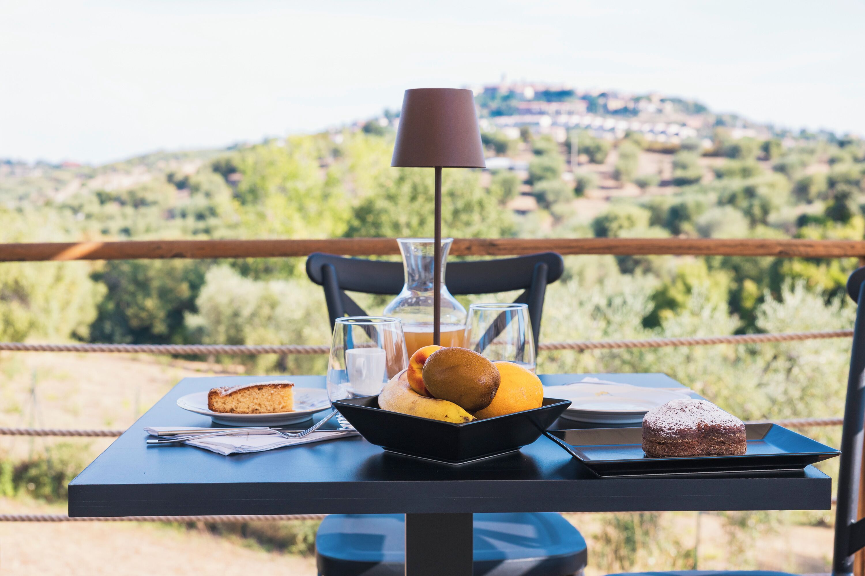 A table with a plate of food, a glass of juice, and a lamp on a balcony overlooking a hilly landscape.