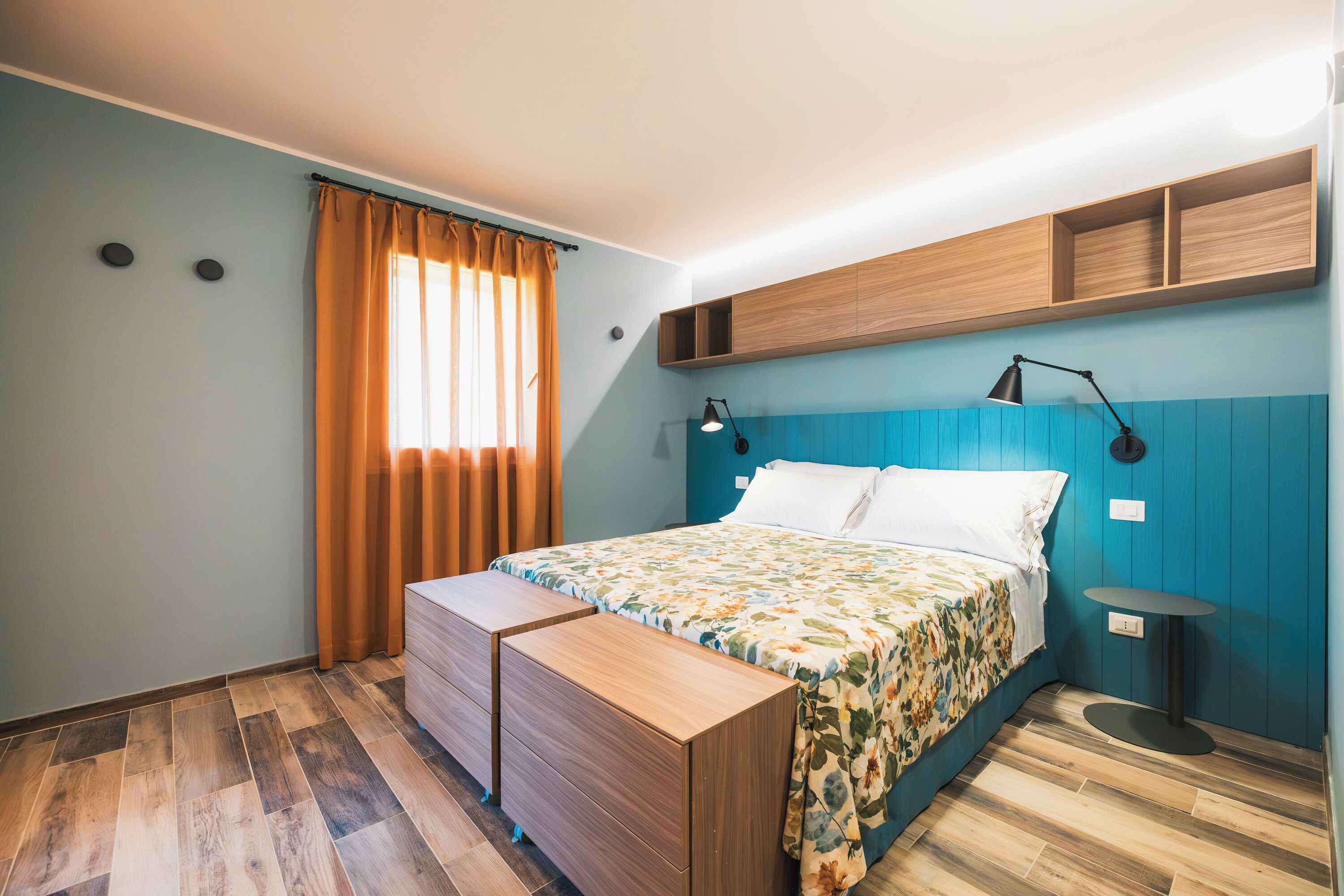 A bedroom with a bed, a desk, a chair, a nightstand, a lamp, and a window with curtains.