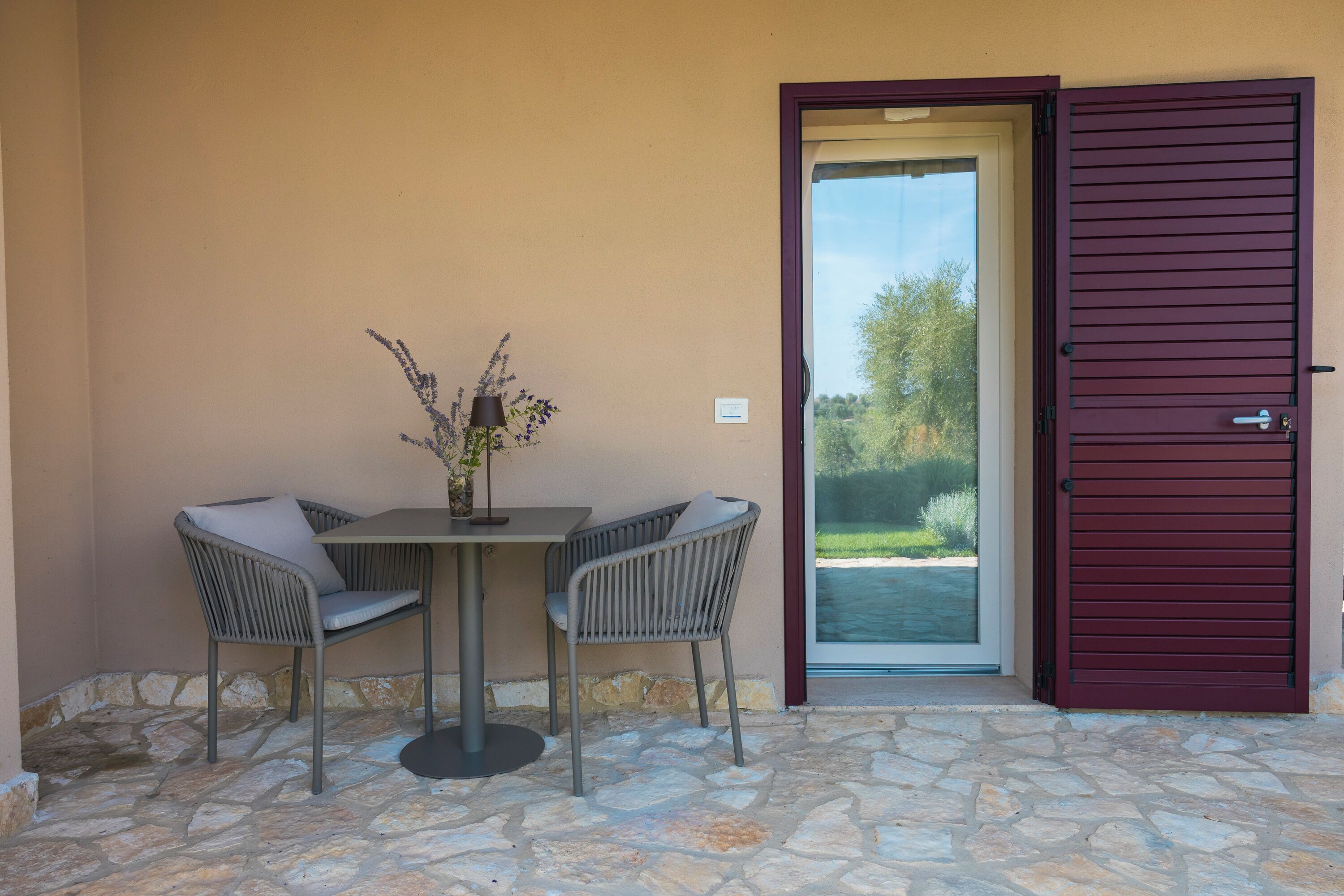An outdoor seating area with two chairs and a table, a potted plant, and a door with a view of the outdoors.
