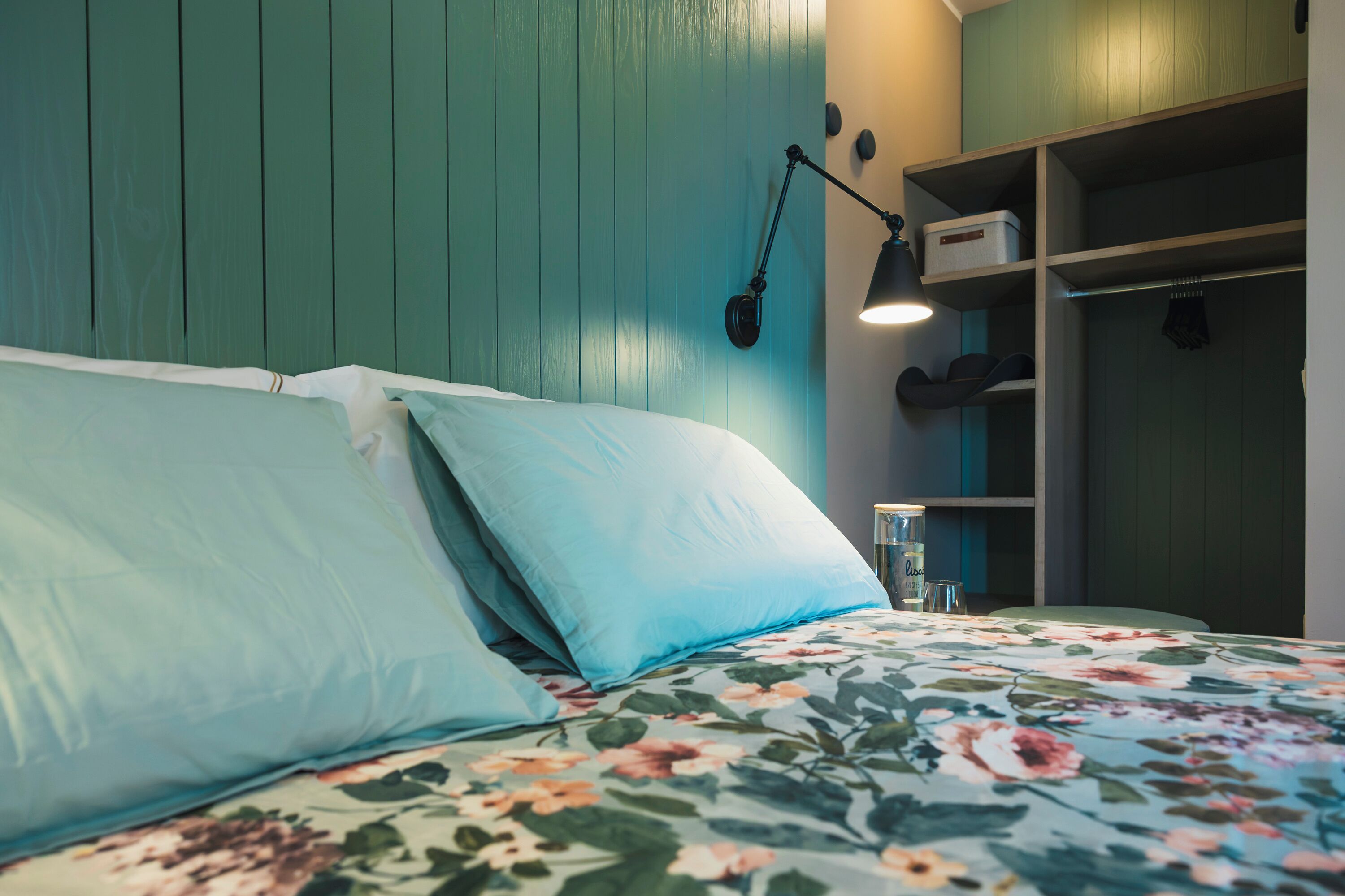 Ein Bett mit floraler Bettwäsche, eine türkisene Wand und ein Regal mit Gegenständen.