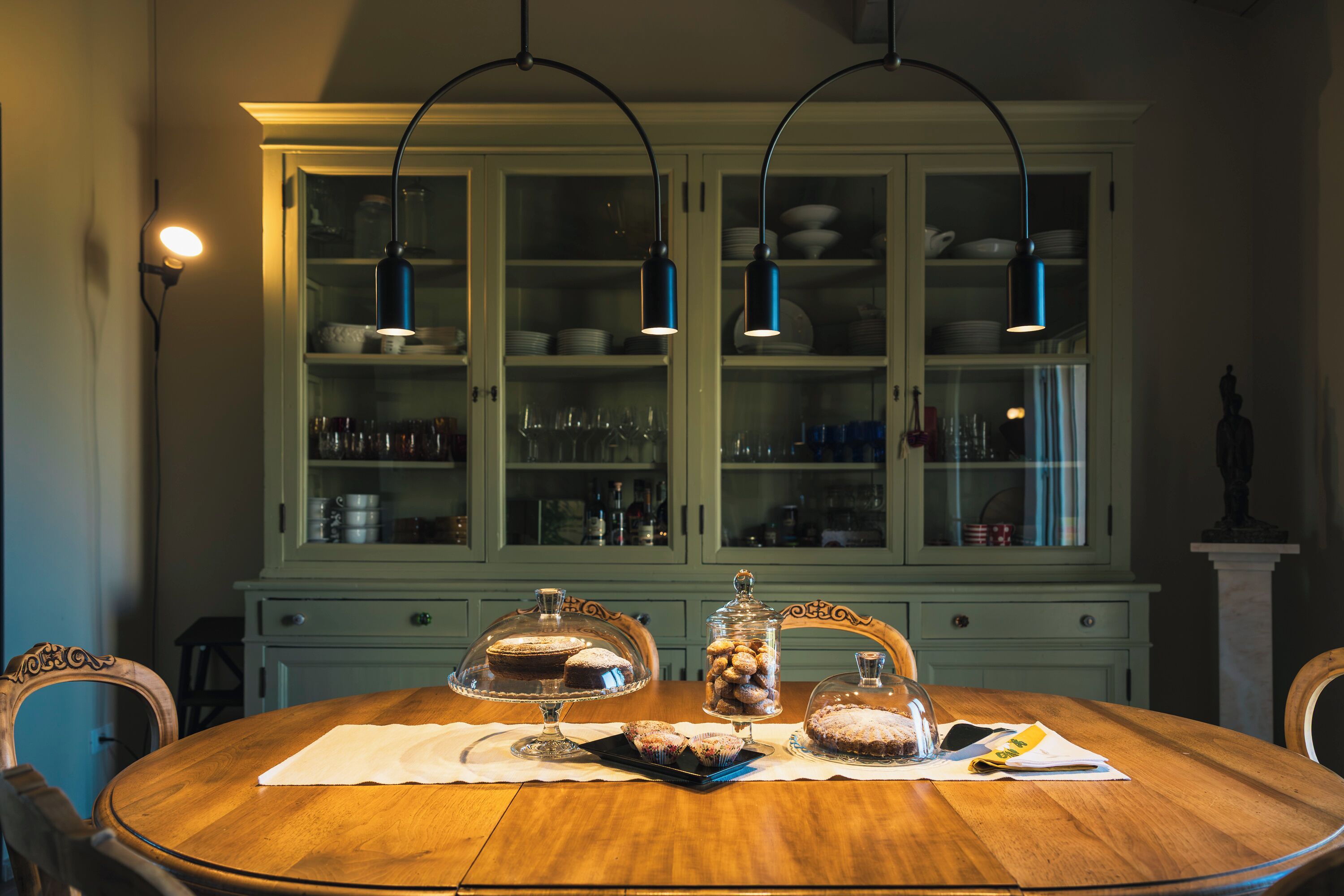A dining table with a wooden finish, surrounded by chairs, in a room with a green cabinet containing glassware and dishes.