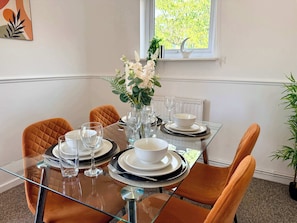 A dining table set with white plates, bowls, and glasses, surrounded by orange chairs, with a vase of flowers and a moon-shaped decoration on the window sill. - 2-Bed Milton Keynes Maisonette Parking (Milton Keynes)