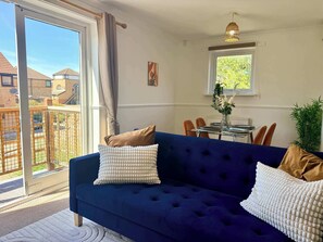 A living room with a blue sofa, two white pillows, a dining table with chairs, a window with a view of buildings, and a hanging light fixture. - Quiet Shenley Church End | Parking & Balcony (Milton Keynes)