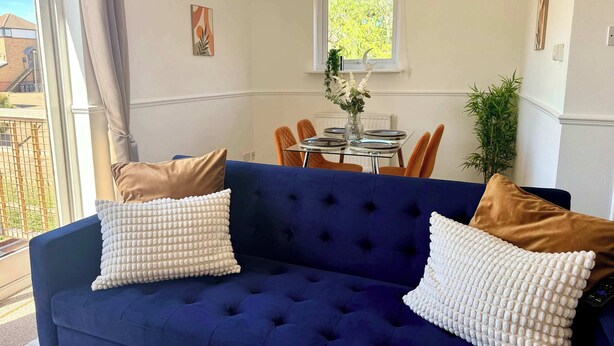 Een blauwe bank met witte en bruine kussens, een eettafel met stoelen, een raam met uitzicht en een hangende lamp.