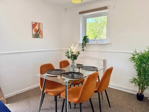 A dining area with a glass table, four orange chairs, a vase with flowers, a window with a view of greenery, and a radiator. - Quiet Shenley Church End | Parking & Balcony (Milton Keynes)