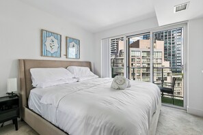 Een slaapkamer met een groot bed, een nachtkastje, een raam met uitzicht op gebouwen en twee ingelijste foto's aan de muur.