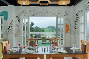 A dining area with a table set for four, a view of a green landscape through open doors, and wall art with leaf patterns. - ELIVAAS Secret Escape Aashiyana 2BHK (Solan)